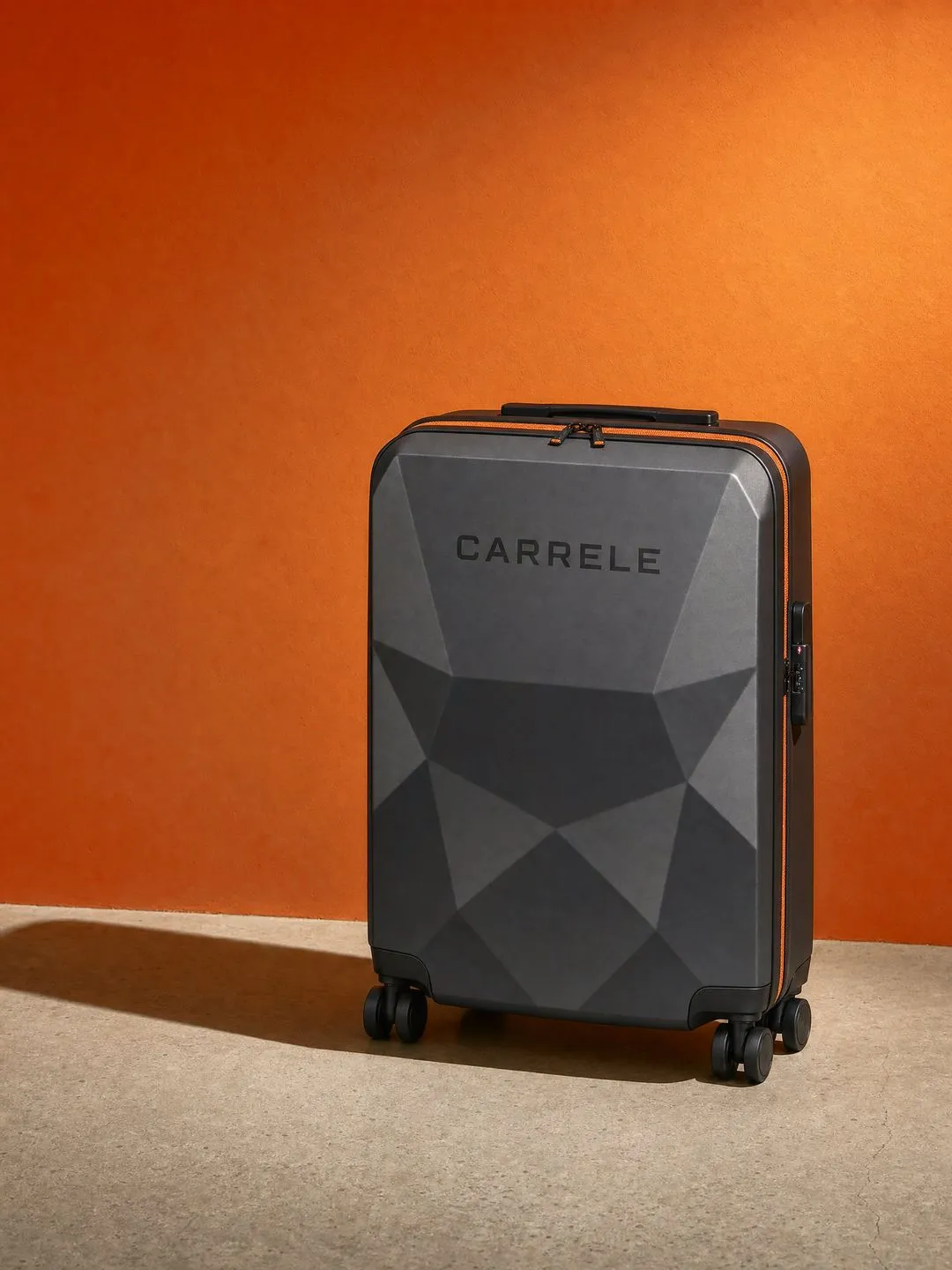 Faceted graphite Carrele suitcase against a bright orange wall.